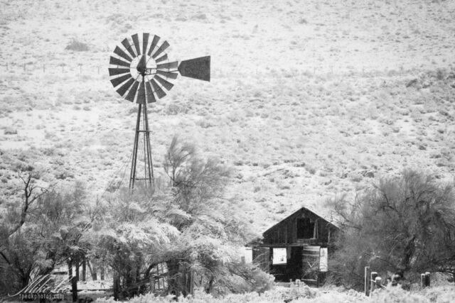 Abandoned Ranch A timeless black and white winter photograph of an abandoned ranch near the old townsite of Amedee in Lassen County, California. A weathered Aermotor windmill towers over a collapsing wooden barn, both dusted with fresh snow from the night before. The surrounding sagebrush and trees glisten under the soft winter light, adding texture and contrast to the quiet rural scene. Once part of a working ranch, the property now stands silent yet dignified against the rolling hills, evoking the endurance and solitude of northern California’s ranching past. A faded “for sale” sign nearby hints at the hope of revival for this forgotten homestead.