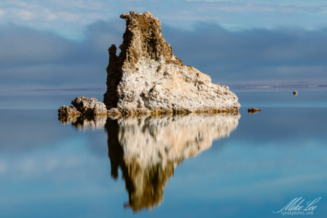 Striking landscape photograph of a large tufa tower at South Tufa on Mono Lake, California, captured on a calm winter day. The towering limestone formation rises from the still, reflective surface of the lake, its intricate texture mirrored perfectly in the glassy water. Subtle fog lingers across the distant shoreline, adding depth and a soft, moody atmosphere beneath a pale blue sky. Formed by the meeting of freshwater springs and alkaline lake water, these iconic tufa structures define Mono Lake’s surreal beauty. This fine art image highlights the geological wonder and tranquil elegance of California’s Eastern Sierra.