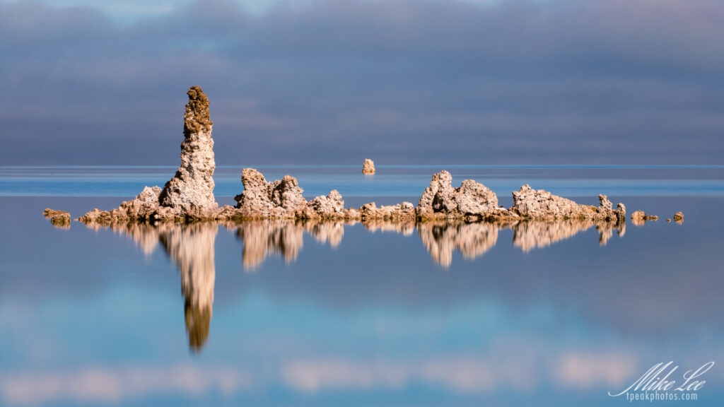 Tranquil fine art photograph of striking tufa formations at South Tufa on Mono Lake, California, captured on a calm winter day. These unique limestone towers—formed when freshwater springs interact with the lake’s alkaline water—rise from the mirror-like surface, their intricate textures perfectly reflected in the stillness below. Soft fog lingers in the background, blending sky and water into a seamless gradient of cool blues and muted earth tones. The composition draws the eye through layers of shape and reflection, highlighting the surreal beauty and geological wonder of Mono Lake’s otherworldly landscape.
