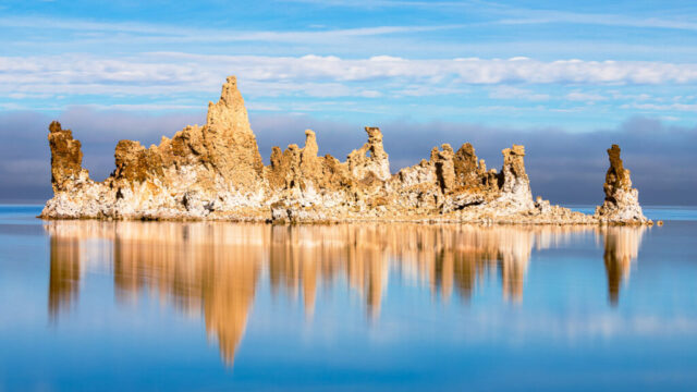 Vibrant landscape photograph of striking tufa formations at South Tufa on Mono Lake, California, captured on a calm winter day. These dramatic limestone spires—created by the interaction of freshwater springs and the lake’s alkaline waters—rise like a natural cathedral from the still, reflective surface below. The long exposure technique smooths the water into a perfect mirror, doubling the intricate textures and warm golden tones of the formations against a backdrop of soft blue sky and lifting fog. This fine art image showcases Mono Lake’s surreal beauty and the timeless elegance of light, geology, and reflection.