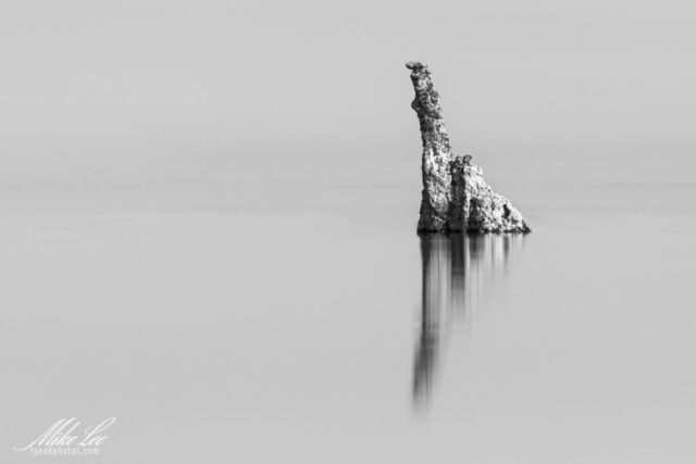 Minimalist black and white fine art photograph of a solitary tufa tower rising from the calm waters of Mono Lake in Mono County, California. Captured with a long exposure and dark neutral density filter, the nearly one-minute shutter smooths the lake into a seamless surface, removing all trace of movement and horizon. The lone limestone formation and its delicate reflection create a striking sense of isolation and balance. This image highlights the intricate textures and geological beauty of Mono Lake’s tufa structures while evoking serenity, simplicity, and timeless stillness.