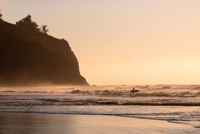 A lone surfer stands in the surf at Trinidad State Beach on California’s North Coast, framed by glowing golden light at sunset. Waves roll toward shore under the warm hues of evening, while sea mist drifts across the water and rugged cliffs rise in silhouette behind. The scene captures the quiet anticipation before entering the ocean, blending the raw beauty of the Pacific with the peaceful glow of coastal twilight.