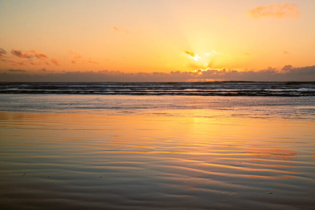 Golden coastal sunset photograph taken at Moonstone Beach in Trinidad, California, where the low tide reveals a wide expanse of wet sand glowing in the evening light. The setting sun slips behind scattered clouds on the horizon, casting radiant beams across the Pacific Ocean and reflecting warm amber tones on the rippled surface of the beach. The flat shoreline and mirrored sand create a peaceful symmetry between sea and sky, while subtle wave lines add texture and depth. This fine art seascape captures the quiet beauty of dusk along California’s rugged North Coast, blending serenity, light, and motion in perfect balance.