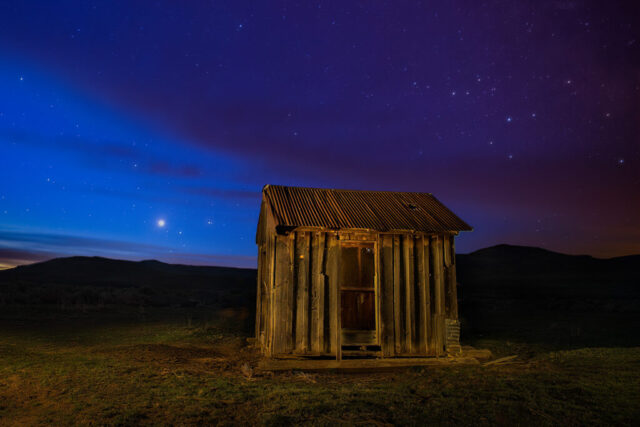 Triple Planet Rise at the Old Shack Pre-dawn astrophotography image titled “Triple Planet Rise at the Old Shack,” captured in the high desert of Lassen County, California. The scene features an abandoned wooden shack with a rusted corrugated roof illuminated by subtle light painting, set beneath a deep twilight sky shifting from violet to indigo. Above the distant ridgeline, Venus, Saturn, and Mars rise in a rare triple alignment, their bright points glowing in the early morning stillness. To the right, the constellation Scorpius arcs into the frame, adding celestial context to the desert landscape. The image was created by stacking multiple high-ISO sky exposures to minimize digital noise and blending them with flashlight-lit frames of the shack, resulting in a vivid and serene composition that unites planetary alignment, rustic history, and the quiet magic of blue hour.