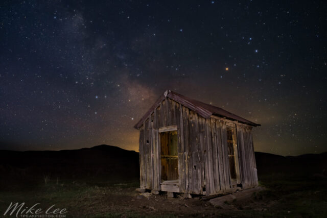 Twighlight at the Old Shack Nightscape photograph titled “Twilight at the Old Shack,” featuring an abandoned wooden shack beneath a twilight sky filled with stars and the faint glow of the Milky Way. Captured in the remote high desert of Lassen County, California, this image shows the weathered structure illuminated with soft, warm light as the horizon fades from gold to indigo. The shack’s rusted tin roof and rough timber walls stand as reminders of the region’s ranching past, likely once serving as a shepherd’s or cowboy line shack. Above, countless stars shimmer in the early morning darkness, with the Milky Way’s core beginning to reveal itself — a quiet meeting of history, solitude, and celestial beauty in the vast desert night.
