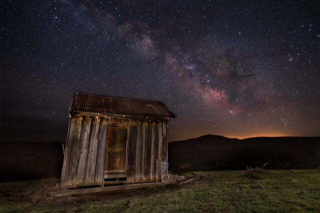 Shack Under the Stars Nightscape photograph titled “Shack Under the Stars,” featuring an abandoned wooden shack illuminated beneath the Milky Way core in the Smoke Creek Desert of Lassen County, California. Captured during the final minutes of astronomical darkness, the image was composed from stacked sky frames to reduce digital noise and several light-painted exposures of the weathered structure. The shack, with its rusted corrugated roof and rough-hewn boards, leans gently into the frame, glowing with soft, warm light against the backdrop of the vast desert night. Above, the Milky Way stretches across the sky in rich tones of violet, gold, and magenta, its galactic dust lanes contrasting sharply with the quiet isolation of the high desert landscape — a timeless meeting of frontier history and cosmic wonder.
