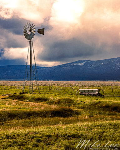 Spring Windmill Vertical A vertical composition highlighting an old Aermotor windmill glowing in warm late-afternoon light against a backdrop of dark storm clouds and the rolling hills of Lassen County, California. The fresh green pasture and irrigation equipment below contrast beautifully with the moody sky above, evoking the changing energy of spring weather in rural northern California ranch country.