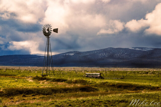 Spring Windmill A dramatic late spring landscape featuring a classic Aermotor windmill standing tall in a lush green pasture under a sky of shifting clouds in Lassen County, California. Captured during unsettled weather, the image contrasts the soft, golden light of the late afternoon sun illuminating the windmill and distant hills with the darker tones of an approaching storm. Just a few miles south, snowfall was blanketing the high country, but here the balance of light and shadow created a perfect rural scene—vivid greens, glowing metal blades, and the timeless feel of open ranchland beneath a moody Sierra sky.