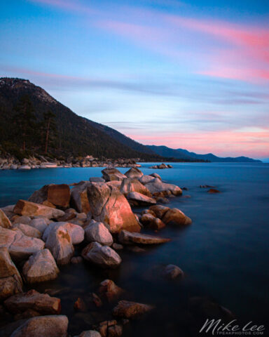 Peaceful long-exposure landscape photograph of Lake Tahoe at dusk, captured from a quiet rocky shoreline along Nevada State Route 28 on the lake’s north side. The two-minute exposure smooths the rippling water into a glasslike surface reflecting the cool blues and soft pink hues of twilight. Rounded granite boulders in the foreground glow with the last warm light of day, leading the viewer’s eye toward forested mountains fading into the distance. This fine art image evokes the calm and solitude of an early summer evening at Lake Tahoe, highlighting the lake’s pristine beauty and the tranquil stillness that follows sunset along the Sierra Nevada.