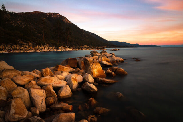 Serene long-exposure panorama of Lake Tahoe at twilight, showing the smooth, glasslike surface of the lake gently meeting the rugged granite shoreline on the Nevada side. Warm tones from the setting sun reflect off the rocks in the foreground, while soft pink and blue streaks paint the sky above the forested Sierra Nevada ridgeline. The wide horizontal composition enhances the sense of calm and openness, capturing the quiet beauty and stillness that settle over Tahoe’s north shore after sunset. A tranquil fine art landscape blending light, color, and texture in perfect alpine harmony.