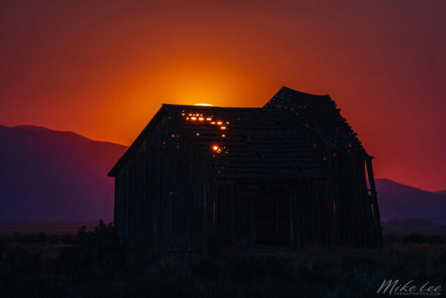 Eerie Glow at the Old Barn A hauntingly beautiful long-exposure photograph of an old, collapsing barn in Lassen County, California, dramatically backlit by the rising Strawberry Moon of 2022. The moon’s orange-red glow, filtered through distant haze and wildfire smoke, radiates through the barn’s broken roof and weathered boards, creating an eerie yet inviting warmth. The surrounding landscape fades into deep purples and shadows, emphasizing the barn’s silhouette and the surreal atmospheric light. This striking image blends decay and beauty, showcasing how even a familiar rural landmark can transform under extraordinary celestial and environmental conditions.
