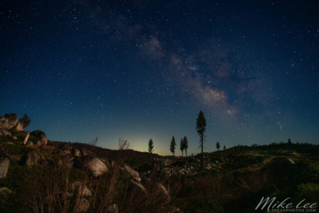 Milky Way over Plumas County on a Full Moon Night Nightscape photograph of the Milky Way arching above a rocky hillside in Plumas County, California, captured on a clear midsummer night illuminated by a full moon. The soft lunar light reveals texture and detail in the boulders, pine trees, and rolling terrain while the galactic core glows faintly but distinctly against the deep blue sky. The combination of bright moonlight and countless stars creates a surreal balance of day and night tones, blending landscape and astrophotography into a single tranquil scene. This image captures the quiet beauty of the Sierra Nevada foothills where celestial wonder meets the rugged calm of Northern California’s high country.