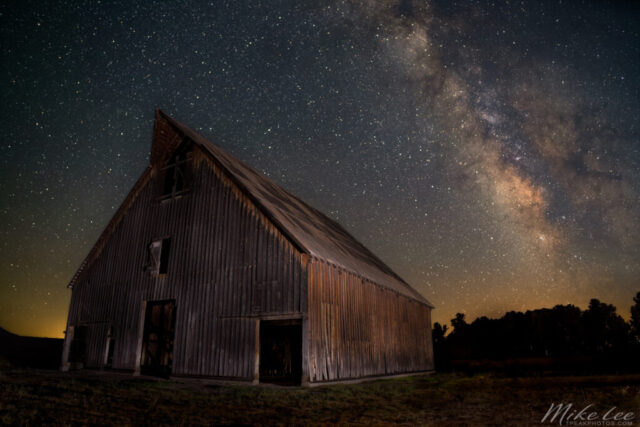 Olsen Barn Nightscape Color version – Nightscape photograph of the historic Olson Barn near Chester, California, illuminated beneath the brilliant core of the Milky Way. The weathered wooden barn, a beloved local landmark set in a wide meadow between the North Fork of the Feather River and Lake Almanor, glows softly under starlight and faint horizon light. Above it, the Milky Way arcs vividly across the sky with rich hues of gold, violet, and deep indigo, revealing dense clusters of stars and dust lanes. This long-exposure image blends local history and cosmic wonder, capturing the peaceful grandeur of the Sierra night sky over this iconic Feather River Land Trust preserve.