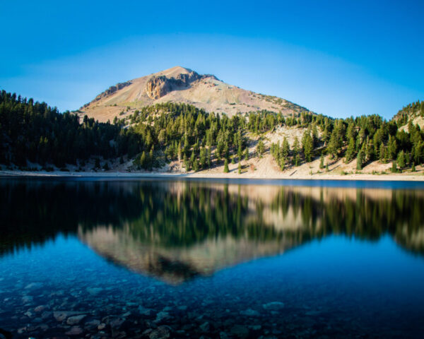 Mount Lassen Reflection Color photograph of Mount Lassen reflected in the still surface of Lake Helen in Lassen Volcanic National Park, California. The late afternoon light illuminates the mountain’s rocky slopes and the dense evergreen forest along the shoreline. A long exposure smooths the wind-ruffled water into a glassy mirror, capturing a nearly perfect reflection of the volcanic peak against a cloudless blue sky. The calm scene contrasts the mountain’s active geothermal nature with the tranquil, high-elevation beauty of the lake.