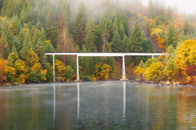 Autumn view of the Lake Britton railroad trestle in Shasta County, California, made famous by the film Stand By Me. The tall white bridge spans calm water surrounded by dense evergreens and brilliant yellow and orange fall foliage under a misty morning sky. Low clouds drift through the forested hillside, creating a tranquil, cinematic atmosphere that captures the moody beauty of Northern California’s Pit River region in autumn.