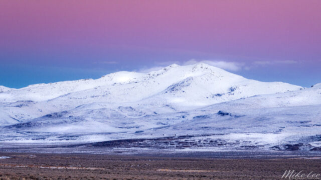 40 North 18 Fahrenheit Minimalist winter landscape photograph of the snow-covered Skedaddle Mountains in Lassen County, California, captured on a frigid morning at 18°F. The scene features soft pastel tones of pink, violet, and icy blue in the dawn sky, blending seamlessly with the white-dusted high desert terrain. Subtle details reveal the faint outlines of munitions bunkers belonging to the historic Sierra Army Depot, nestled within the stark expanse. This fine art image conveys the quiet isolation and cold beauty of the Great Basin landscape, where the frozen stillness of winter morning light transforms an austere desert mountain range into a serene, dreamlike vista.
