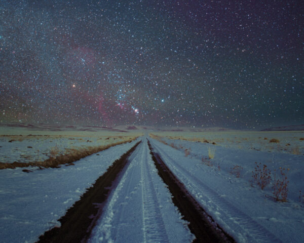 Cold Lonely Road to Orion Astrophotography image of a snow-covered rural road stretching into the horizon beneath a vast winter night sky rich with stars and nebulae in the Orion constellation. Captured using a series of stacked long-exposure frames to minimize digital noise, the photo reveals a striking mix of natural airglow and hydrogen-alpha emission. A soft greenish haze of atmospheric nightglow glimmers across the upper sky, while deep red and magenta tones from Orion’s nebulae shine vividly thanks to an astro-modified camera with its infrared-blocking filter removed. The snow’s cool tones contrast beautifully with the colorful celestial expanse, creating a serene and otherworldly winter landscape that highlights the quiet intersection of Earth and the cosmos.