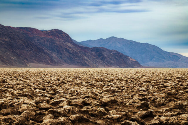 Expansive view of the Devil’s Golf Course in Death Valley National Park, California, where jagged salt formations stretch endlessly beneath a soft winter sky. This alien-like terrain—formed by evaporated ancient lakebeds and sculpted by relentless wind—creates a surreal, textured landscape that contrasts dramatically with the muted tones of the surrounding Panamint Mountains. Captured from the edge of the salt flat on a calm January afternoon, the image highlights the raw beauty and desolate power of one of Earth’s most extreme environments. Perfect for lovers of desert landscapes, geology, and fine art nature photography that celebrates the extraordinary artistry of the natural world.