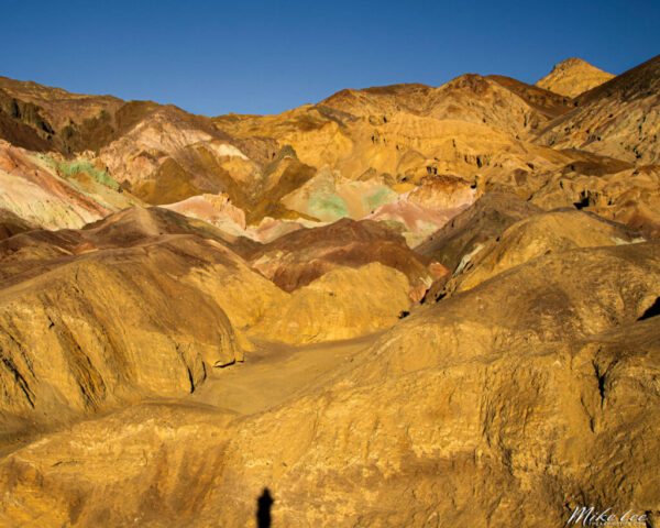 Vivid mineral colors sweep across the sculpted hillsides of Artist’s Palette in Death Valley National Park, California. Captured in the warm, low light of late afternoon, the scene reveals brilliant shades of gold, red, pink, and green created by the oxidation of volcanic minerals over millennia. The rugged texture of the landscape glows under a cloudless desert sky, embodying the surreal beauty and geological complexity that make this location one of Death Valley’s most iconic viewpoints. A small human shadow offers a sense of scale amid the grandeur—an artistic nod to the connection between nature and observer.