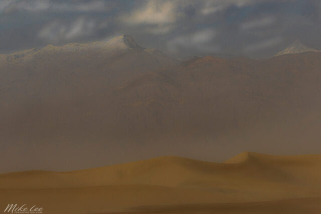 Golden dunes swirl beneath a haze of windblown dust at the Mesquite Flat Sand Dunes in Death Valley National Park, California. Captured with a telephoto lens on a stormy winter morning, this image compresses the sweeping desert foreground against the distant, snow-capped Panamint Range. The airborne dust softens the light and mutes the rugged tones of the mountains, creating a painterly, atmospheric effect that feels almost surreal. The contrast between the warm sand and cool high peaks speaks to the extremes that define Death Valley—a landscape of both beauty and brutality. A captivating fine art photograph for lovers of desert light, dramatic weather, and the vastness of the American Southwest.