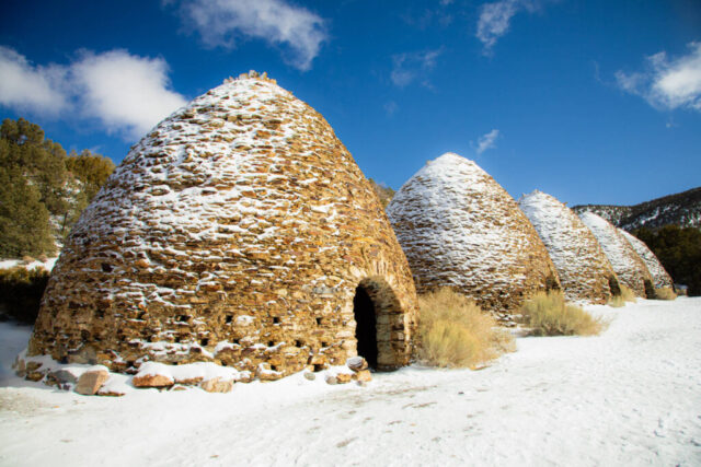 Photograph of the Wildrose Charcoal Kilns in Death Valley National Park, California, taken on a crisp winter morning after a light snowfall. The ten conical, beehive-shaped kilns—built from native stone and mortar in 1877 to supply charcoal for nearby silver and lead smelters—stand in a row beneath a vivid blue sky at roughly 6,800 feet in the Panamint Mountains. Their weathered stone surfaces are lightly capped with snow, creating a striking contrast of warm earth tones against the white ground and deep sky. This image captures both the rugged beauty of the desert highlands and the enduring legacy of California’s mining history, a rare winter view of one of the best-preserved examples of 19th-century industrial architecture in the American West.