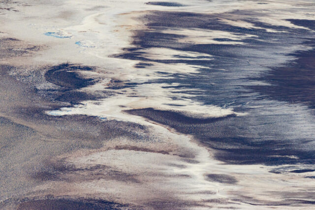 Aerial landscape photograph of Badwater Basin in Death Valley National Park, California, viewed from the high vantage point of Dante’s View at sunset. The image captures intricate abstract patterns formed by crystalline salt and mineral deposits across the basin floor, illuminated by the soft, golden light of late afternoon. Subtle waves of texture and tone—ranging from pale whites and warm beiges to cool blues and purples—create a painterly, almost otherworldly effect that reflects the constant transformation of this extreme desert environment. Shaped by wind, water, and time, the salt flats reveal the geological artistry of one of North America’s most unique landscapes. A compelling minimalist study in light, texture, and natural abstraction, perfect for lovers of desert photography and modern fine art.