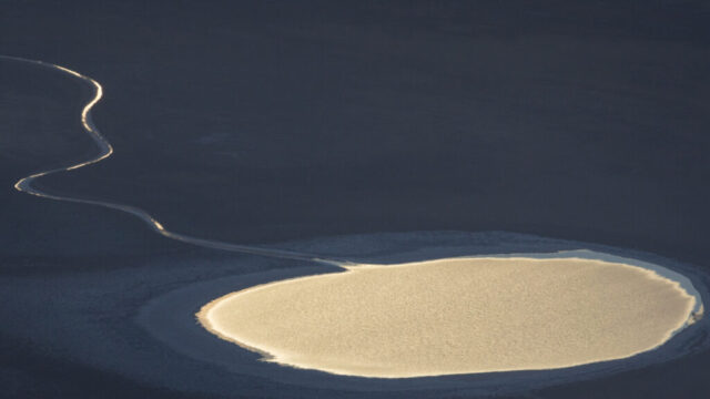 Aerial telephoto view of Badwater Basin in Death Valley National Park, California, captured from Dante’s View as the late afternoon light faded on a January 2023 day. The photograph highlights a pale, circular salt formation surrounded by darker salt flats, with a faintly glowing meandering ridge of salt leading toward it—an illusion that at first glance resembles a reflective pool of water. The contrast between the bright, clean salt and the surrounding darker crust suggests recent ponding from floods that occurred in the summer of 2022, now dried and crystallized under the desert sun. The warm light grazing the surface emphasizes the subtle textures and abstract geometry of this vast basin, evoking both natural minimalism and the surreal beauty of Death Valley’s ever-changing landscape.