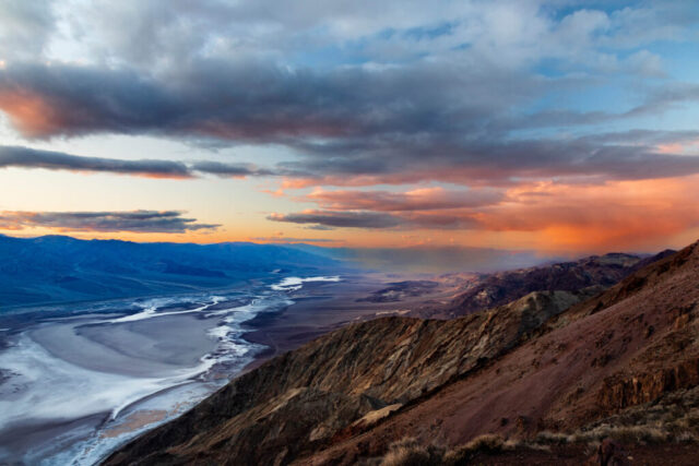 Panoramic view of Death Valley National Park captured from Dante’s View during a vivid winter sunset. The photograph looks down from over 5,600 feet of elevation across the vast expanse of Badwater Basin, where the white salt flats wind through the valley floor nearly 300 feet below sea level. Warm golden and fiery orange light from the setting sun illuminates the Panamint Range to the west, contrasting with the deep blues and purples of the distant mountains and clouds. The rugged foreground ridgeline adds texture and scale, emphasizing the immense vertical relief of this dramatic desert landscape. A striking image that captures the raw beauty, contrast, and atmospheric color of Death Valley at day’s end.