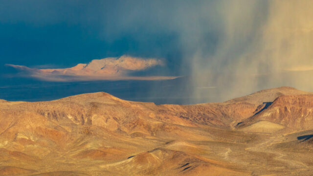 Atmospheric desert landscape photograph taken in Death Valley National Park, California, showing a golden range of sunlit hills in the foreground beneath a dramatic sky filled with shifting rain squalls. Captured on a January afternoon from near Dante’s View, the image reveals a striking contrast between the warm, ochre tones of the rugged terrain and the cool, moody blues of the distant mountains and storm clouds. A veil of mist and rain drifts across the scene, partially obscuring the background and adding a sense of depth and motion. The interplay of light and shadow evokes the ever-changing moods of the desert, where fleeting weather patterns transform the arid landscape into something ethereal and alive.