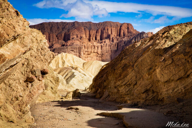 Photograph of Red Cathedral as seen from within Golden Canyon in Death Valley National Park, California. The image showcases the vibrant red rock cliffs of the cathedral formation rising dramatically above the lighter golden and beige badlands in the foreground. Sunlight filters through high clouds, softening the midday light and accentuating the rich textures and layered geology of the canyon walls. The narrow, winding path leads the viewer’s eye toward the towering rock face, revealing the striking contrast between the warm sandstone tones and the deep blue sky. A vivid depiction of Death Valley’s sculpted desert terrain, highlighting the beauty, isolation, and geological wonder of this iconic hiking destination.