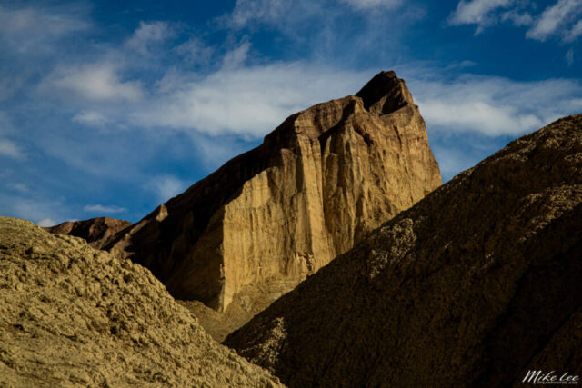 Landscape photograph of Manly Beacon rising above the rugged terrain of Golden Canyon in Death Valley National Park, California. The towering, sharply defined rock formation glows in warm desert tones under a soft canopy of high clouds, which diffuse the sunlight to reveal intricate textures and layered sedimentary details. The composition emphasizes the beacon’s dramatic, angular peak as it stands out against the vivid blue sky, framed by the darker canyon walls below. Captured on a calm January afternoon, this image highlights the timeless beauty and geological grandeur of one of Death Valley’s most iconic landmarks—a testament to the power of erosion, sunlight, and time in shaping the American desert landscape.