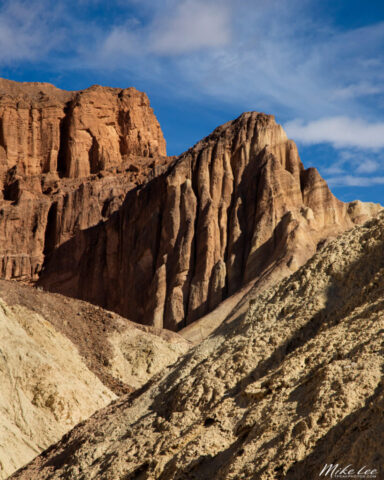 Photograph looking up Golden Canyon in Death Valley National Park, California, showcasing the dramatic formations of Red Cathedral and the surrounding golden badlands. The towering rust-red cliffs rise above lighter, weathered ridges in the foreground, their layered textures illuminated by soft midday light filtering through high, wispy clouds. Subtle shadows and warm highlights reveal the intricate geology and erosion patterns that define this iconic desert landscape. The composition draws the viewer’s eye toward the jagged peaks beneath a vivid blue sky, capturing the timeless grandeur and color contrast that make Golden Canyon one of Death Valley’s most photogenic and geologically fascinating destinations.