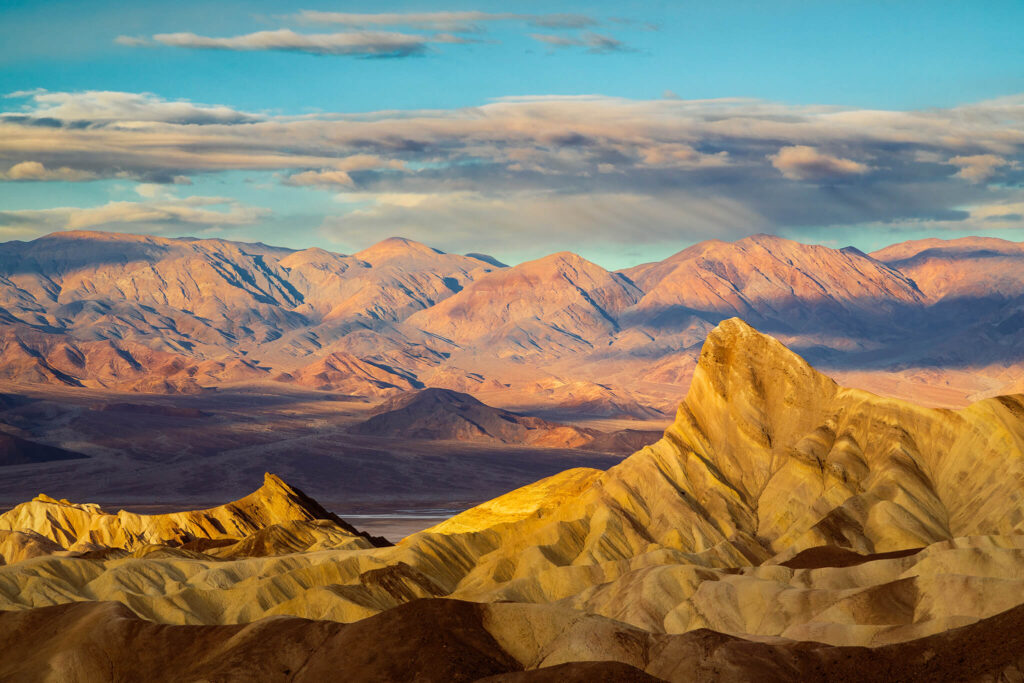 Morning light over Zabriskie Point in Death Valley National Park, California, illuminating the sculpted badlands and the prominent peak of Manly Beacon. The golden rays of sunrise wash across the layered ridges and eroded formations, revealing rich tones of yellow, amber, and sienna against the cool purples and blues of the distant Panamint Mountains. Soft clouds stretch across the sky, adding depth and contrast to the scene while emphasizing the vastness of this ancient landscape formed from sedimentary deposits of Furnace Creek Lake. A timeless view that captures the quiet grandeur and geological artistry of one of Death Valley’s most iconic and photographed locations.