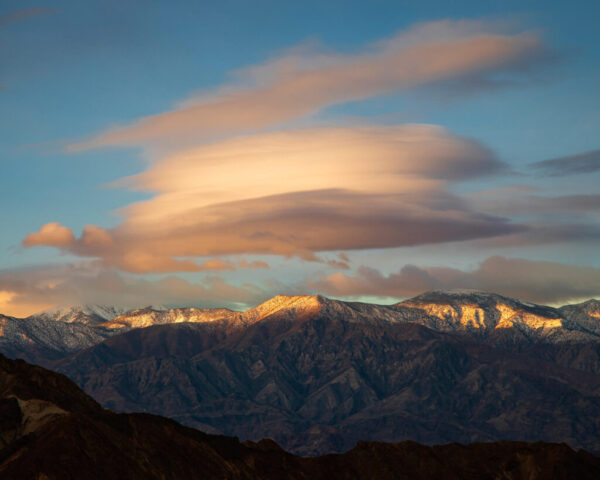 Photograph of a glowing lenticular cloud hovering above the snow-dusted peaks of the Panamint Range in Death Valley National Park, California. Captured on a calm January morning, the image shows the cloud illuminated in soft golden light as the rising sun breaks through an earlier layer of low clouds. The rugged mountain ridgeline, shaped by tectonic forces, contrasts beautifully with the smooth, layered form of the lenticular cloud—its edges tinted with warm hues against a serene blue sky. This rare and atmospheric scene highlights the interplay of weather, light, and geology that defines the dramatic landscapes of the California desert.