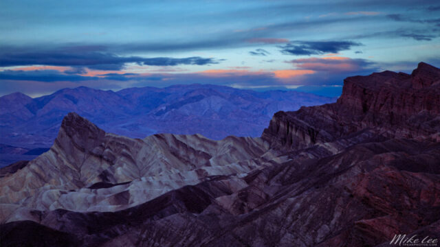 Pre-dawn landscape at Zabriskie Point in Death Valley National Park, California, captured as the cool tones of blue hour transition toward the warm glow of sunrise. The photograph features the dramatic textures and layered formations of the badlands, with Manly Beacon rising prominently on the left and the rugged red cliffs of Red Cathedral on the right. Subtle light from the approaching dawn highlights the striped ridges and deep valleys, while soft pink and purple hues streak the sky above the distant Panamint Mountains. This serene moment reveals the timeless beauty and geological artistry of one of Death Valley’s most iconic and beloved viewpoints.