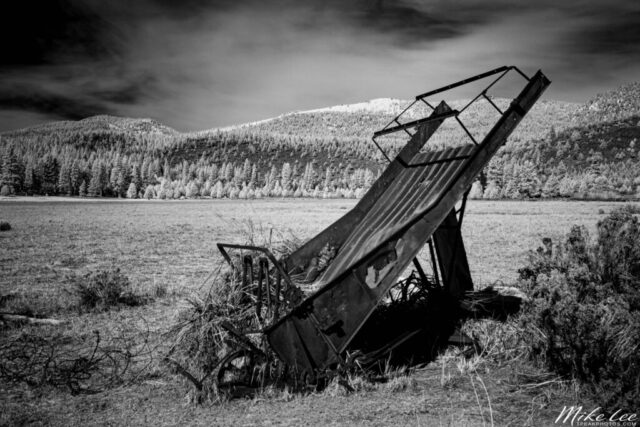Rusting Hay Elevator in Infrared A moody black and white infrared photograph of a rusting hay elevator leaning in a sunlit field at Ranch Park near Susanville, California. Captured with infrared light, the image transforms the rural landscape into a striking high-contrast scene where the metallic textures of the abandoned farm equipment contrast sharply against the glowing trees and pale grasses of the surrounding meadow. Snow-dusted hills rise in the background under a deep, dark sky, enhancing the timeless and weathered feel of this piece. This evocative image celebrates Susanville’s ranching heritage and the quiet beauty of forgotten agricultural relics in northern California’s high country.