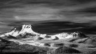 Black and white infrared photograph of Mount Lassen and the Chaos Crags in Northern California, viewed from Hat Creek Rim near Old Station. The snow-covered lava dome of Lassen Peak rises sharply against a dark, textured sky with streaking high clouds. To the right, the jagged peaks of Chaos Crags catch the light, their forms emphasized by strong contrast and deep tonal range. In the foreground, a forested landscape spreads across rolling terrain, its trees rendered in bright white tones typical of infrared imagery. The dramatic lighting and contrast evoke a timeless, Ansel Adams–like aesthetic.