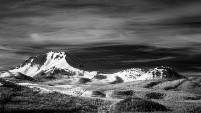 Mount Lassen and Chaos Crags in Invisible Light Black and white infrared photograph of Mount Lassen and the Chaos Crags in Northern California, viewed from Hat Creek Rim near Old Station. The snow-covered lava dome of Lassen Peak rises sharply against a dark, textured sky with streaking high clouds. To the right, the jagged peaks of Chaos Crags catch the light, their forms emphasized by strong contrast and deep tonal range. In the foreground, a forested landscape spreads across rolling terrain, its trees rendered in bright white tones typical of infrared imagery. The dramatic lighting and contrast evoke a timeless, Ansel Adams–like aesthetic.