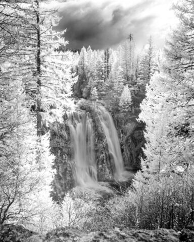 The 8th Wonder Infrared Infrared black and white photograph of Burney Falls in McArthur-Burney Falls Memorial State Park, Shasta County, California, captured with a full-spectrum camera and an 830nm deep infrared filter. The surreal tonal range transforms this iconic waterfall into a dreamlike scene, where the cascading water glows softly between bright, luminescent trees and dark volcanic cliffs. Dramatic spring clouds add texture and depth to the sky, enhancing the ethereal contrast typical of infrared imagery. This unique fine art interpretation offers a fresh perspective on one of Northern California’s most photographed landmarks, blending natural grandeur with otherworldly light and atmosphere.
