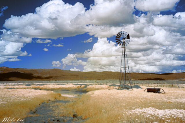 Rural Aermotor False Color A surreal infrared landscape featuring a classic Aermotor windmill standing tall against a backdrop of billowing cumulus clouds near Madeline, California. Captured with a full spectrum modified camera and a 590nm near-infrared cut filter, the image blends visible yellows, oranges, and reds with infrared light to create striking false colors. The dry ranchland and reflective pond below the windmill contrast beautifully with the deep sky tones produced by channel swapping in post-processing. This remote Lassen County scene along US-395 embodies the rugged quiet of rural northern California, where cattle ranching and open skies define the landscape.