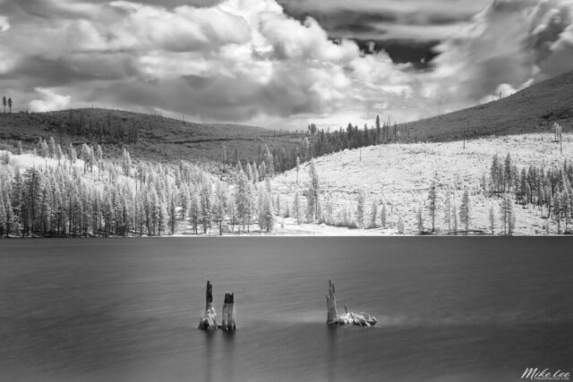 Striking long exposure infrared photograph of Blue Lake in Lassen County, California, captured using an 830-nanometer deep infrared filter. The surreal monochrome tones render evergreens and hillsides in glowing white while the lake’s smooth, glassy surface contrasts against textured clouds drifting overhead. The skeletal remains of old tree stumps rise from the water, adding visual depth and timeless character to the composition. Taken on a calm June afternoon, this fine art infrared landscape reveals the quiet isolation and dreamlike beauty of one of Northern California’s most peaceful and remote mountain lakes.