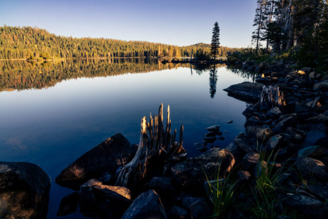 Serene morning landscape photograph of Gold Lake in Plumas County, California — the largest body of water in the scenic Lakes Basin region. Captured from the rocky shoreline near Gold Lake Campground, this image reflects the calm stillness of dawn as the golden light of sunrise brushes the trees and distant hills. The mirror-like surface of the lake perfectly echoes the surrounding conifers and soft summer sky, creating a balanced composition of warmth and tranquility. A peaceful tribute to Northern California’s high-country beauty, ideal for those who find solace in nature’s quiet moments.