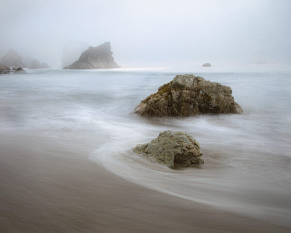 Soft fog drifts over sea stacks and shoreline rocks at Harris Beach State Park in Brookings, Oregon, creating a calm and ethereal coastal mood. The gentle motion of retreating waves blurs into misty textures, while sunlight faintly filters through the fog in the distance, bathing the scene in soft, silvery light. Captured at the southern end of the park’s southern beach, this minimalist seascape embodies the essence of the Pacific Northwest coast—quiet, mysterious, and ever-changing. Perfect for coastal decor and ocean lovers drawn to the tranquil beauty of foggy Oregon beaches and the soothing rhythm of the sea.