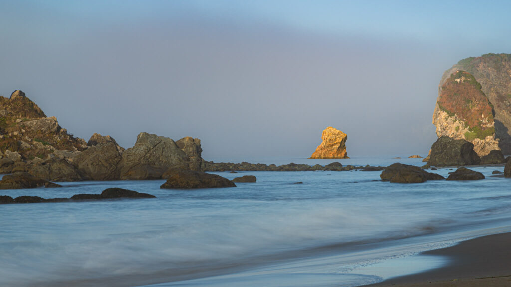 Golden morning light illuminates a rugged sea stack and rocky shoreline at Harris Beach State Park in Brookings, Oregon, moments before fast-moving fog rolled in from the Pacific. The tranquil surf glides across the wet sand while soft mist begins to veil the cliffs and sea stacks in the distance. This fleeting moment of clarity captures the essence of Oregon’s coast—where sun and fog dance in constant motion, creating ever-changing light and atmosphere. A serene coastal landscape perfect for lovers of the Pacific Northwest, seascape photography, and the quiet magic of golden hour along Oregon’s wild southern shoreline.