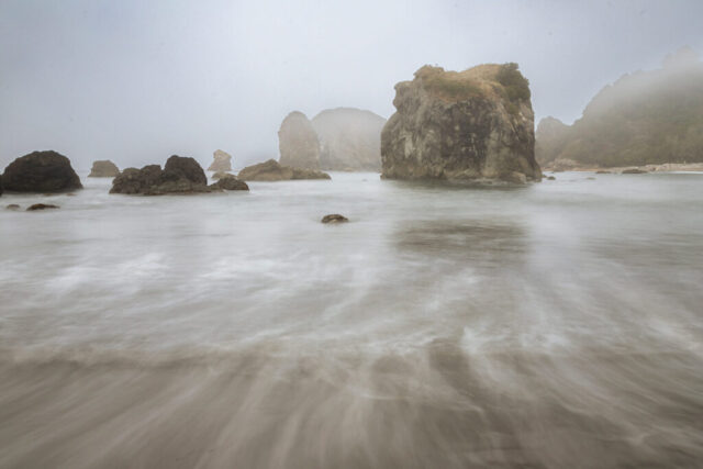 Fog drapes the rugged sea stacks of Harris Beach State Park in Brookings, Oregon, as soft waves sweep across the sand in graceful, blurred motion. Captured on a misty October morning with a slow shutter speed, the receding surf forms elegant leading lines that draw the eye toward the layered rock formations rising through the coastal haze. This atmospheric seascape captures the tranquil, moody beauty of Oregon’s southern coast—where fog, tide, and stone meet in perfect harmony. Ideal for coastal decor, ocean lovers, and those captivated by the serene mystique of the Pacific Northwest shoreline.