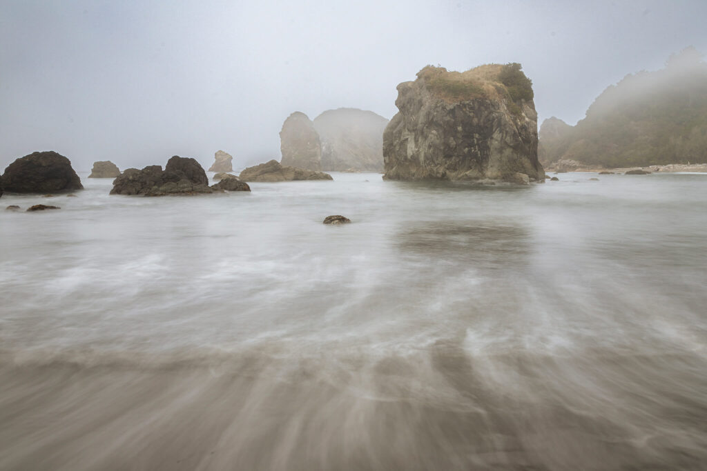 Fog drapes the rugged sea stacks of Harris Beach State Park in Brookings, Oregon, as soft waves sweep across the sand in graceful, blurred motion. Captured on a misty October morning with a slow shutter speed, the receding surf forms elegant leading lines that draw the eye toward the layered rock formations rising through the coastal haze. This atmospheric seascape captures the tranquil, moody beauty of Oregon’s southern coast—where fog, tide, and stone meet in perfect harmony. Ideal for coastal decor, ocean lovers, and those captivated by the serene mystique of the Pacific Northwest shoreline.
