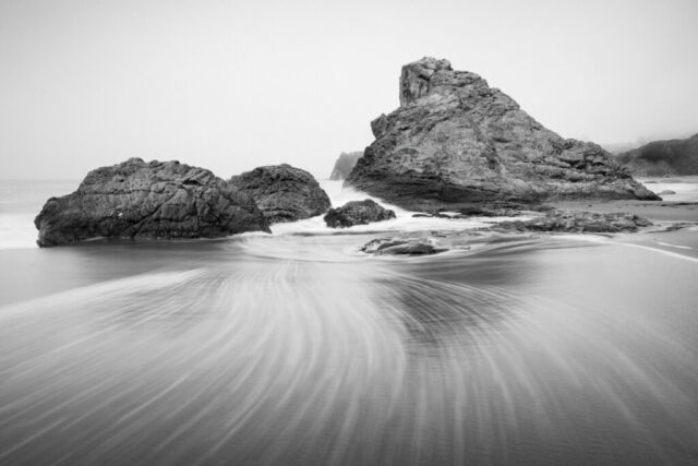 A long-exposure black-and-white seascape at Harris Beach State Park in Brookings, Oregon, captures the motion of receding waves swirling gracefully toward a rugged sea stack. Shot on a hazy afternoon with about a one-and-a-half-second shutter speed, the blurred water forms elegant striations that create a natural leading line through the composition. The monochrome treatment emphasizes texture and contrast, highlighting the interplay between smooth, flowing surf and the rough volcanic rock formations. This minimalist coastal photograph embodies the raw power and tranquility of the Oregon coast—perfect for fine art collections, coastal-inspired interiors, and lovers of moody seascape photography.