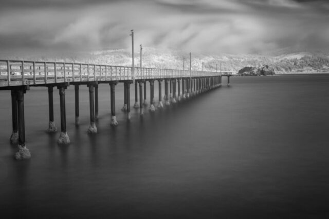 A long-exposure black-and-white infrared photograph of the Crescent City Boardwalk in Northern California, rendered with ethereal tones and striking contrast. Captured with a full-spectrum modified camera and 830-nanometer IR filter, the two-minute exposure smooths the Pacific waters into a glassy surface, emphasizing the boardwalk’s elegant structure and symmetry. The historic jetty stretches toward the distant hills of Del Norte County, softly illuminated beneath shifting coastal light. Subtle details—like gulls perched atop lampposts—add a quiet sense of life to this timeless seascape. A refined fine art image that blends maritime history with infrared minimalism, perfect for coastal interiors and collectors of long-exposure photography.
