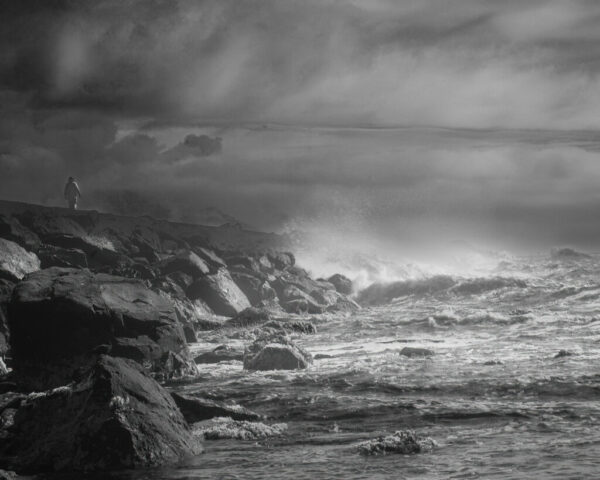 Black and white deep infrared photograph of the jetty between Battery Point and Crescent Harbor in Crescent City, California. Captured with an 830nm filter on a full spectrum modified camera, the image shows a solitary person walking along the rocky breakwater as waves crash violently against it under stormy skies. The glowing infrared tones emphasize texture, contrast, and atmosphere, creating a dramatic seascape that blends human scale with the raw power of the Pacific coast.
