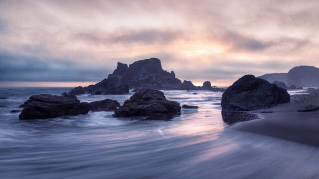 Soft pastel light filters through drifting coastal fog in this late afternoon seascape from Harris Beach State Park in Brookings, Oregon. The scene captures the fleeting glow that appeared roughly half an hour before sunset, with gentle tones of lavender, peach, and blue reflecting across the receding waves. Jagged sea stacks rise from the shoreline, their dark silhouettes contrasting beautifully with the glowing sky and smooth motion of the surf. This tranquil moment, photographed on a quiet October evening, embodies the serene and moody beauty of the Oregon coast—perfect for ocean lovers, landscape photography enthusiasts, and those drawn to the peaceful rhythm of coastal twilight.